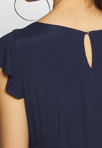 Takana näkymä henkilöstä, joka on pukeutunut tummansiniiseen mekkoon, jossa on liehuvat hihat ja pieni avainreikäpainike kauluksessa.