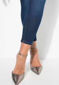 Metaliczne szare szpilki z wyciągniętym noskiem i paskami na kostkach, noszone z ciemnoniebieskimi dżinsami, które są lekko skrócone na kostkach, z widocznym przeszyciem.