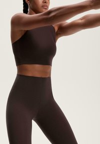 Braunes sportliches Cropped-Top mit einem einseitigen Schulterträger, kombiniert mit hochgeschnittenen Leggings, beide aus einem glatten, elastischen Stoff.