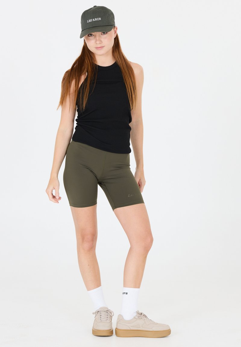 Top sans manches noir côtelé, shorts ajustés vert olive, baskets beige avec semelles tan, et chaussettes blanches mi-mollet. La casquette affiche le texte "LES ARGS".