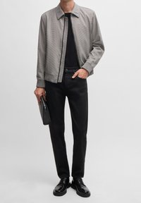 Chaqueta gris claro con cremallera y un diseño texturizado, camiseta negra ajustada, pantalones negros y zapatos negros brillantes. Sosteniendo un clutch negro.
