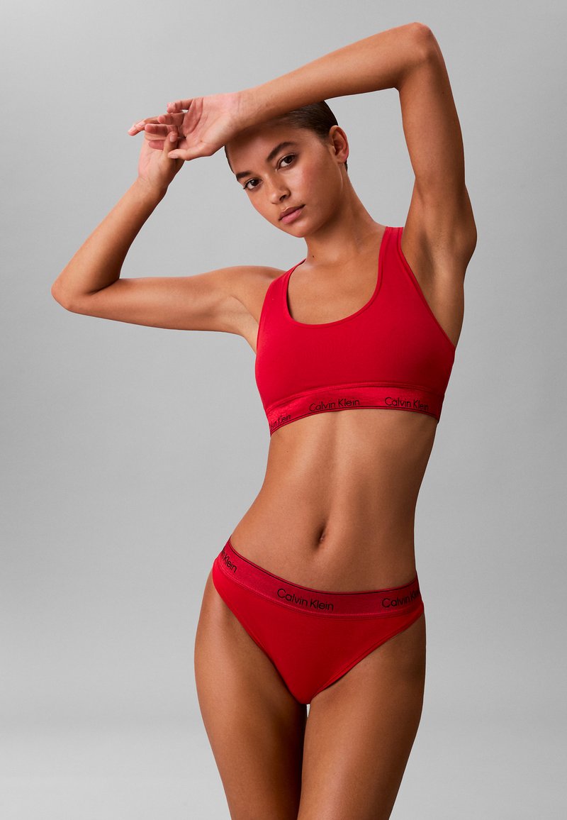 Set di bralette e bikini in cotone rosso con cintura in logo Calvin Klein, caratterizzato da una texture liscia e un design aderente. Lo sfondo è grigio.