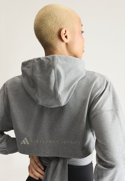 Person with short blonde hair wearing a gray cropped hoodie with hood up and Stella McCartney logo on the back, hands on hips.