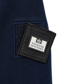 Čierno-biela tkaná nášivka s korunou a textom "Weekend Offender" pripevnená na tmavomodrom materiáli na suchý zips.
