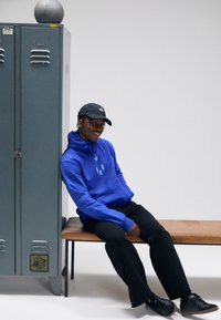 Sweatshirt azul com capuz e design de logótipo, calças pretas, sentado num banco de madeira ao lado de um cacifo cinzento, usando um boné preto e sapatos pretos.