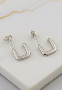 Orecchini a cerchio in argento con forma quadrata, texture liscia e finitura lucida, dotati di chiusura a post. Posati su una superficie di marmo chiaro.