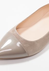 Chaussure plate en daim beige avec un bout pointu en cuir laqué taupe brillant. Texture lisse avec un design sans couture et des accents minimalistes.