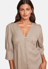 Beige linendress med dyp V-hals, pufferm og dekorative knapper nedover fronten. Glatt tekstur, enkel og elegant design.