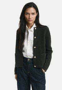 Kvinna med långt mörkt hår som bär en svart knäppt kofta över en vit skjorta och mörkblå jeans, stående med en hand i fickan.