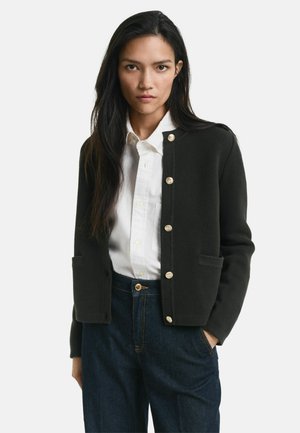 Femme avec de longs cheveux noirs portant un cardigan noir à boutons sur une chemise blanche et un jean bleu foncé, se tenant avec une main dans la poche.