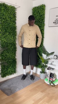 Homem está em um ambiente fechado sobre um tapete cinza, vestindo uma jaqueta bege, calções pretos, meias brancas e sapatos pretos, cercado por plantas verdes e equipamento fotográfico.