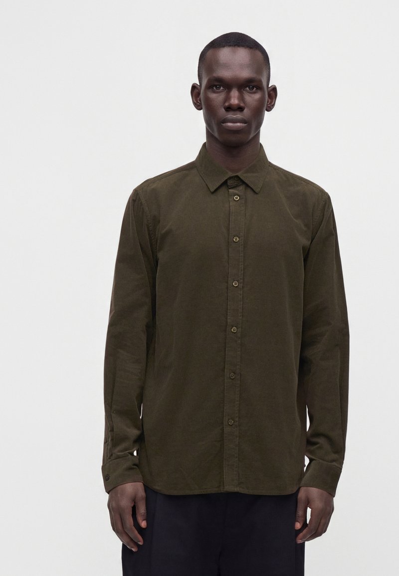 Homme debout, face à l'avant, portant une chemise boutonnée à manches longues vert olive et un pantalon sombre sur un fond clair uni.