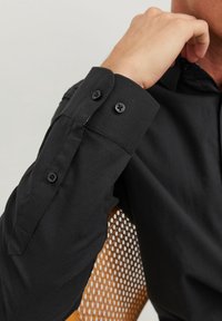 Svart långärmad skjorta med knäppta manschetter, som har en mjuk textur och ren design, visad mot en vävd stol som bakgrund.