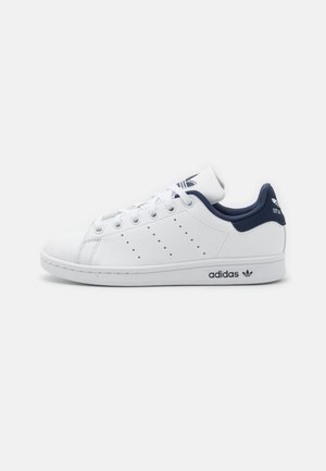 Baskets blanches Adidas Stan Smith avec languette de talon bleu marine, lacets, bandes perforées et logo noir sur la semelle et le talon.