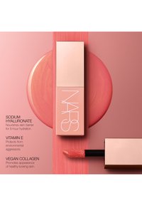 Lipgloss in een slanke, rechthoekige, zachtroze tube met een glanzende afwerking. Bevat een zachte applicator met product erop, gezet op een roze achtergrond.