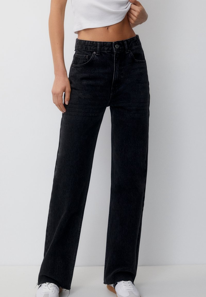 Black high-waisted jeans with wide legs, featuring a button closure, five pockets, and a slight textured finish. Worn with a fitted white top.