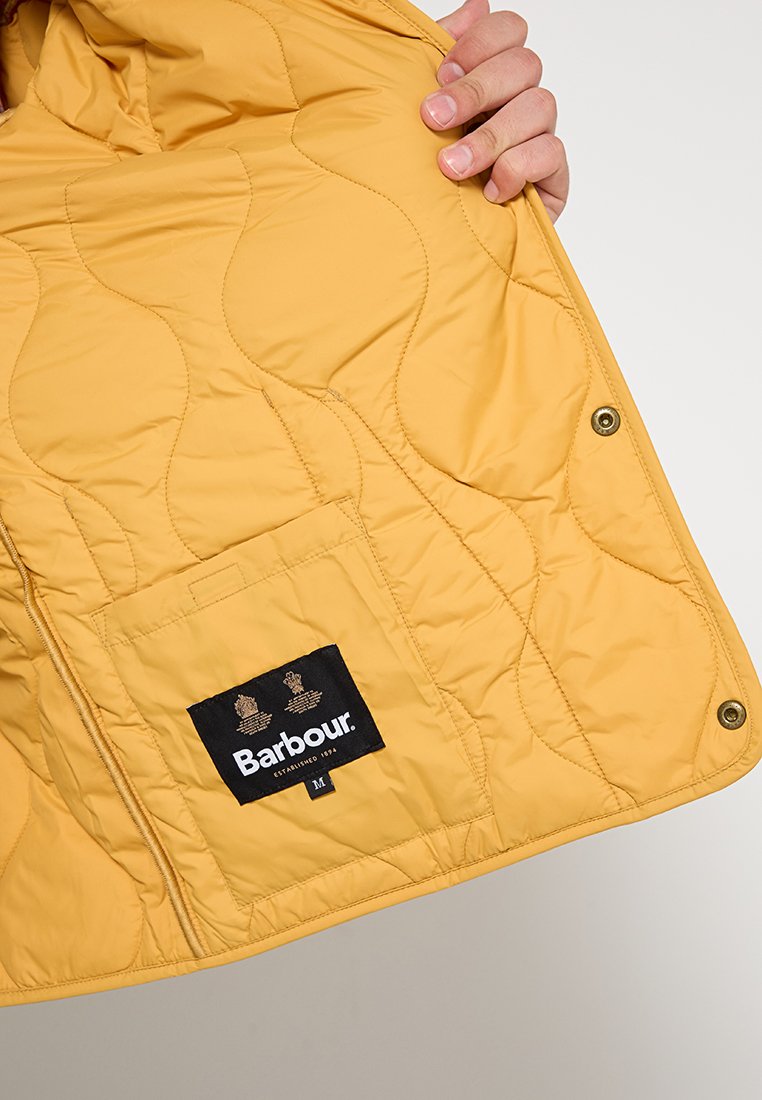 Eine Hand hält eine senfgelbe, gesteppten Jacke offen, die eine Innentasche mit einem schwarzen Barbour-Label und Druckknöpfen zeigt.