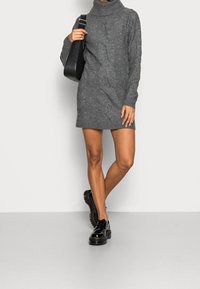 Une personne portant une robe à col roulé en tricot grise, tenant un sac à épaule noir et des chaussures à plateforme noires et épaisses, se tenant devant un fond uni.