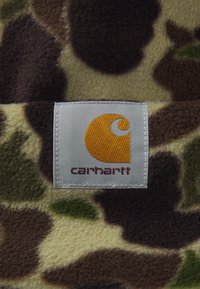 Tejido de forro polar con un patrón de camuflaje en tonos marrón, verde y beige; incluye una etiqueta blanca con un logo amarillo y texto rojo.