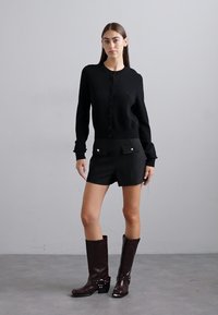Cardigan nero con bottoni, pantaloni corti neri su misura con tasche a patta e stivali di pelle marrone al ginocchio con hardware decorativo.