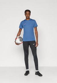 Μπλε αθλητικό μπλουζάκι κοντού μανικιού με ροζ λογότυπο Nike, συνδυασμένο με εφαρμοστά μαύρα παντελόνια με υφασμάτινες λεπτομέρειες. Κρατώντας μια σχεδιασμένη μπάλα ποδοσφαίρου.