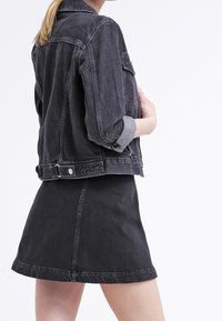 Kobieta w czarnej dżinsowej kurtce z podwiniętymi rękawami i pasującą czarną dżinsową spódnicy, stojąca na tle jednolitego białego tła.