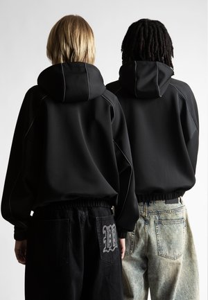 Divi cilvēki stāv ar muguru pret kameru, ģērbtā melnos kapucēs; viens valkā melnas bikses ar baltu izšuvumu, otrs - gaiši zilo džinsu bikses.