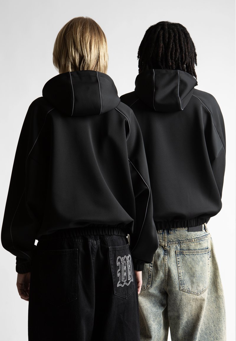 Divi cilvēki stāv ar muguru pret kameru, ģērbtā melnos kapucēs; viens valkā melnas bikses ar baltu izšuvumu, otrs - gaiši zilo džinsu bikses.