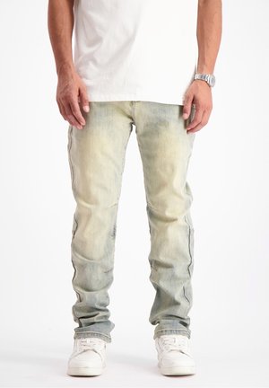 Hombre con vaqueros ajustados de lavado claro, zapatillas blancas, camiseta blanca y reloj de pulsera plateado, de pie sobre un fondo blanco.