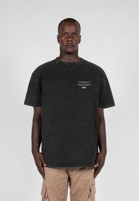 Sort oversized t-shirt i en falmet vask med korte ærmer. Har små hvide bogstaver på forsiden. Modellen bærer beige cargo-bukser.