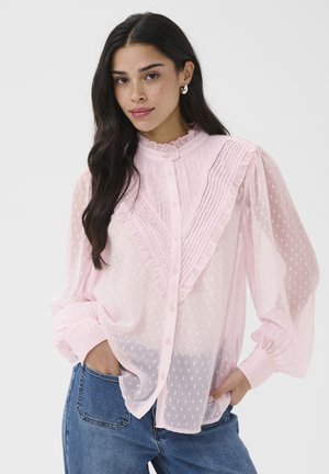 Hosszú sötét hajú nő átlátszó rózsaszín fodros blúzban, melyen texturált pöttyök vannak, kék farmerral, kezét a zsebébe dugva.