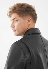 Zwarte leren jas met een scherpe kraag en een slank ontwerp, met zichtbare stiksels en een gladde, matte textuur.