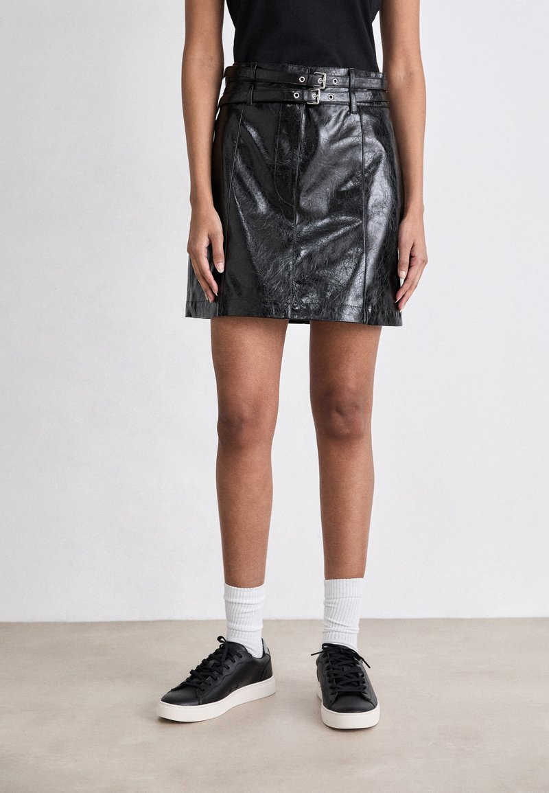 Personne portant une jupe noire en simili cuir avec ceinture à double boucle, baskets noires à semelles blanches, chaussettes blanches et un haut noir.