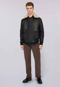 Chaqueta bomber de cuero negra con cuello de borrego beige, dos bolsillos frontales, combinada con un suéter gris oscuro y pantalones marrones. Zapatos negros.