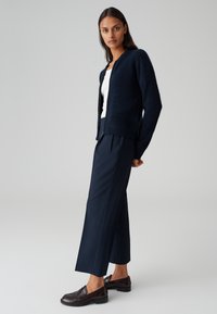 Cardigan intenso in navy con scollo rotondo, abbinato a pantaloni navy su misura e mocassini scuri, su uno sfondo neutro.