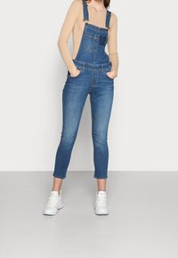 Jeanslatzhosen mit verstellbaren Trägern, einer Vordertasche und einem schmal zulaufenden Bein. Farbe: mittleres Blau mit verwaschenen Stellen.