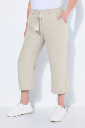 Femme portant un pantalon beige raccourci avec cordon et des baskets blanches, les mains dans les poches, debout devant un fond uni.