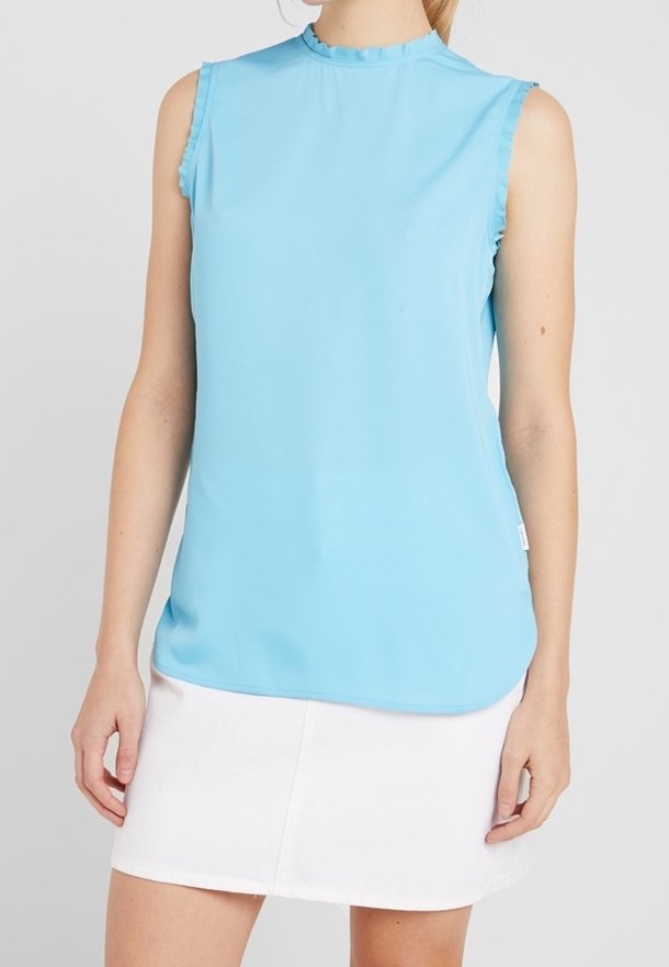 Femme portant un haut sans manches bleu clair aux bords froncés, associé à une jupe blanche, sur un fond gris uni.