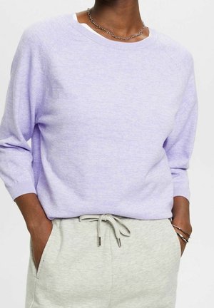 Personne portant un pull violet clair à manches longues, un pantalon gris clair à cordon, un collier chaîne argenté et des bracelets argentés avec les mains dans les poches.