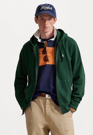 Jonge man met een blauwe Ralph-hoed, groene zip-hoodie, gestreepte rugbytrui, beige broek en een zonnebril die aan zijn shirtkragen hangt.