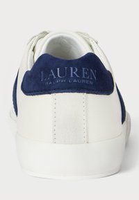 Biele kožené tenisky s námorníckymi semišovými akcentmi na päte, ktoré majú vystúpené modré písmená "LAUREN RALPH LAUREN" a textúrovaný povrch.