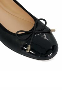 Black ballet flat with a glossy patent toe cap, rounded shape, simple bow detail, and gold-tone hardware accents. Soft inner lining.