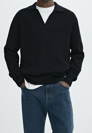 Homme portant un pull en maille noir par-dessus un t-shirt blanc avec un jean bleu, les mains dans les poches, sur un fond clair uni.