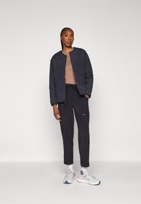 Gequiltte zwarte bomberjack met een ronde hals, een bruin shirt, donkere broek en grijze sneakers. Jack heeft een gestructureerd patroon.