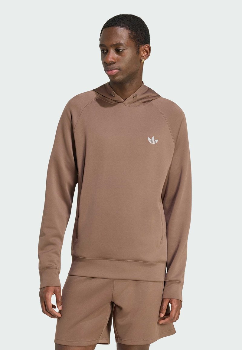 Homme portant un sweat à capuche Adidas marron et un short assorti, debout les mains détendues, regardant légèrement vers le bas et la gauche.