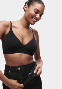 Glunderende vrouw met haar in een paardenstaart draagt een zwarte bralette en maakt een hoge taille zwarte denim rok los voor een effen achtergrond.