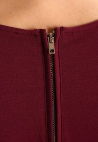 Burgundský přiléhavý top se středovým kovovým zipem, se hladkým povrchem a krátkými rukávy. Úhledné švy podél okrajů.