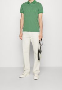 Polo vert avec un logo de marque discret, associé à un pantalon crème et des baskets blanches. Tenant un sac noir avec une bandoulière.