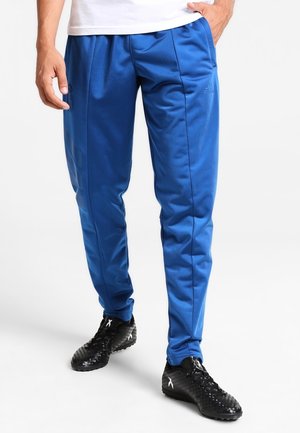 Mężczyzna w niebieskich dresach Adidas i czarnych korkach stoi z jedną ręką w kieszeni na białym tle.