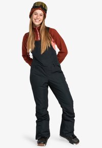 Overoles negros de nieve con bolsillos laterales, usados sobre una camiseta de manga larga roja. El modelo lleva botas de nieve negras y un gorro de lana marrón con gafas.